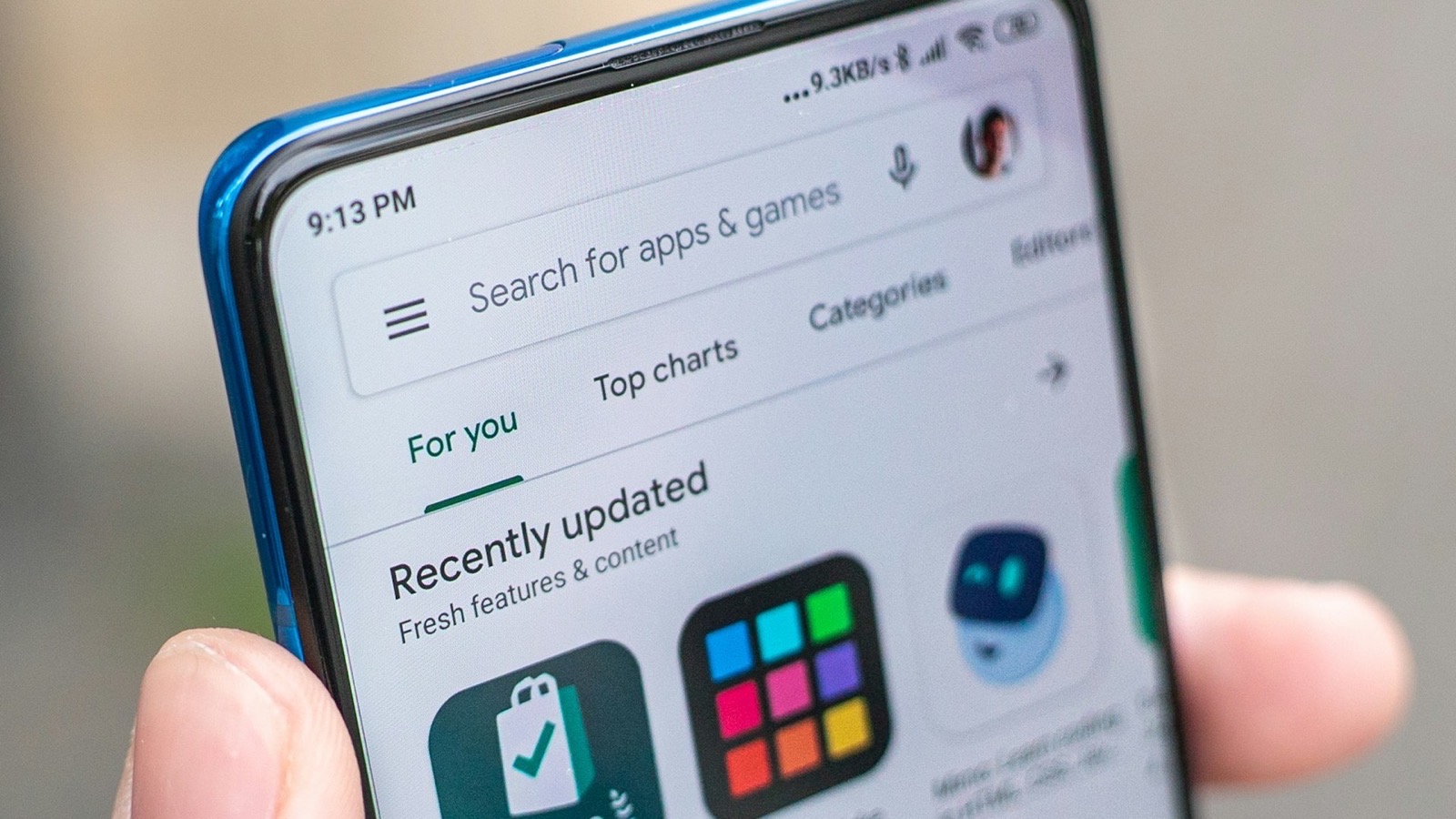 A hand holding an Android phone with the Google Play Store app open, showing app icons under Recently updated
