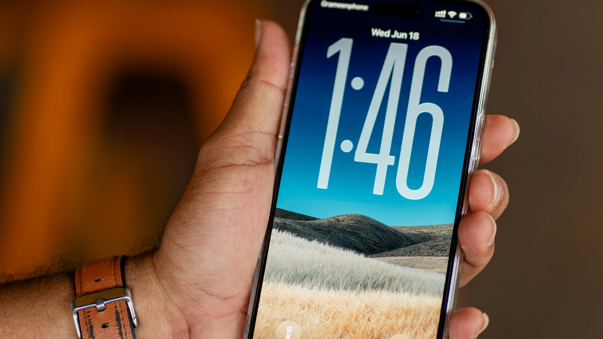 A close-up of a hand holding an iPhone with its lock screen lit up, showing the time against a warm blurred background