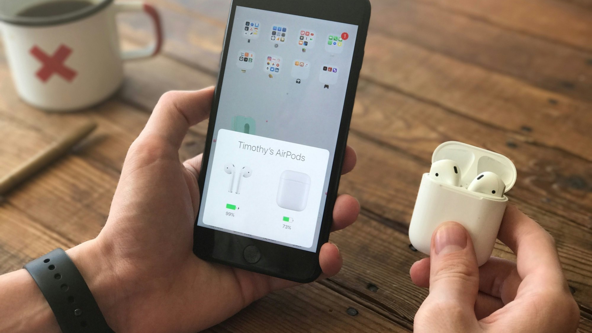 A hand holding an iPhone with its screen lit up, next to an open AirPods case on a wooden desk