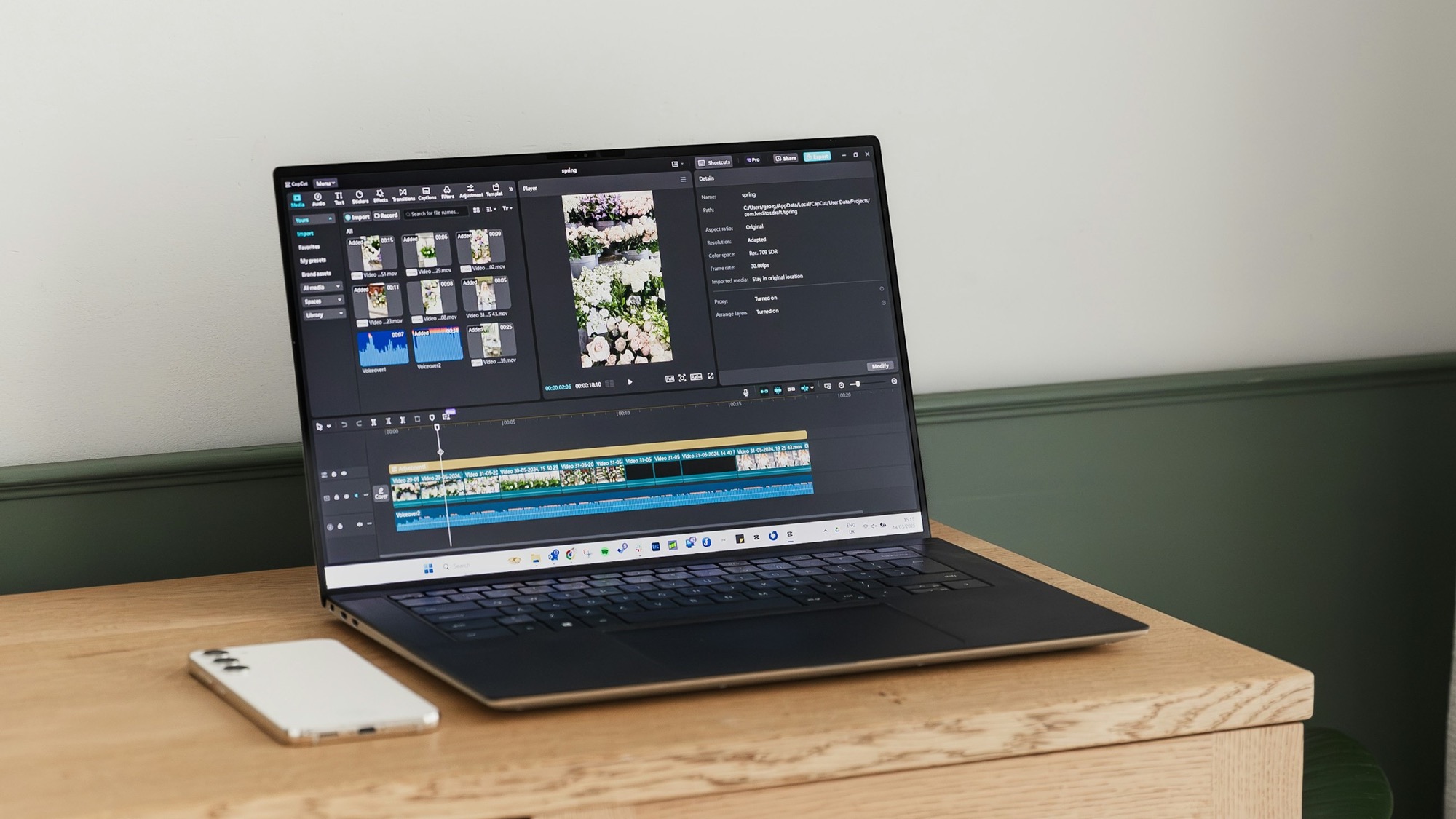 A laptop open on a wooden desk, showing a video editing timeline with clips, audio waveforms, and a preview panel