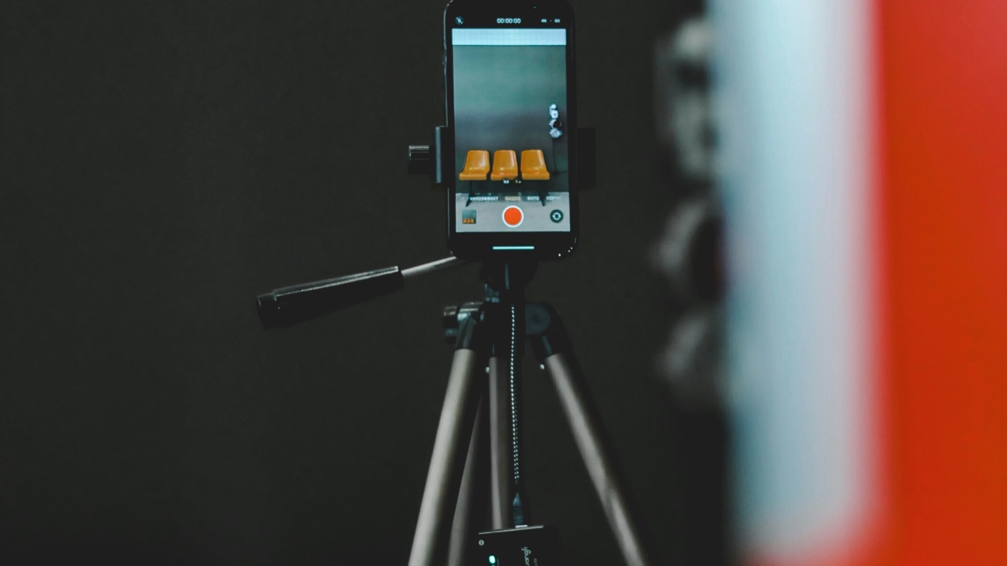 A smartphone mounted on a tripod, screen lit up and recording, in a dim studio setting